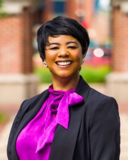 Headshot of a smiling Black woman with short hair wearing a bright purple top and black suit jacket