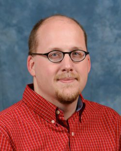 Headshot of smiling white man with short hair and a goatee and glasses in a red checked shirt