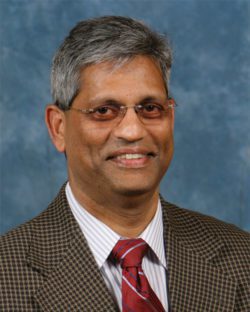 Headshot of smiling Indian man with gray hair and glasses wearing a checked suit and tie