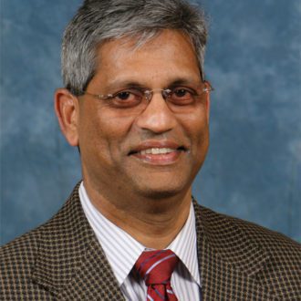 Headshot of smiling Indian man with gray hair and glasses wearing a checked suit and tie