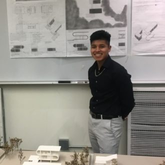 A male Judson student poses in front of a poster with a building design while a 3-D model of the building rests on a table in front of him