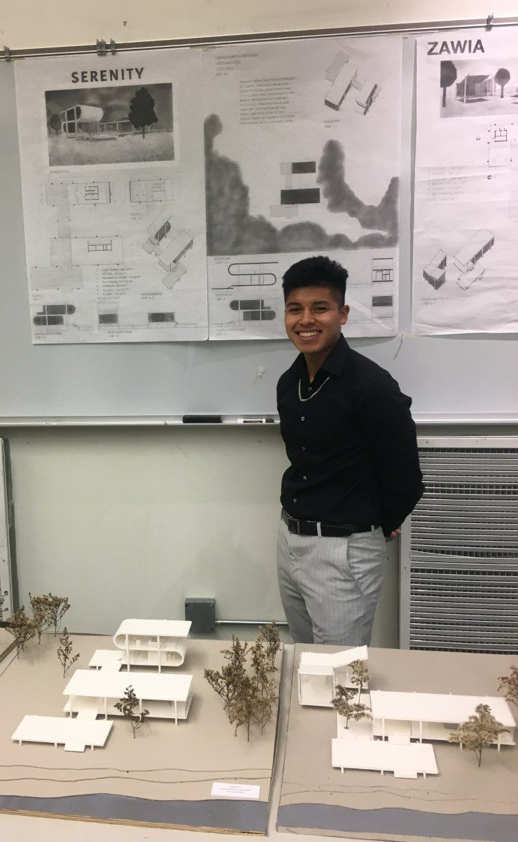 A male Judson student poses in front of a poster with a building design while a 3-D model of the building rests on a table in front of him