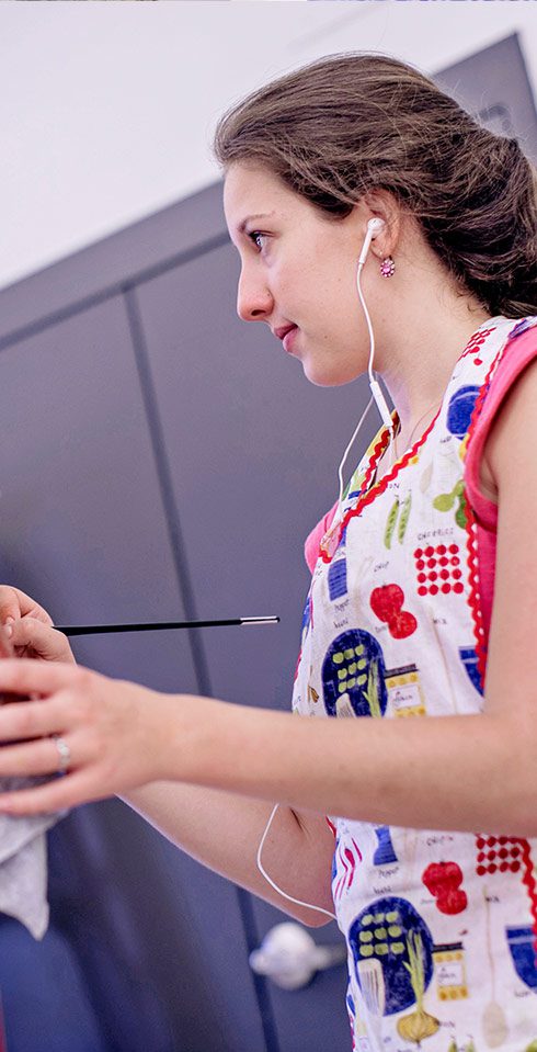 A female Judson student wearing earbuds looks across the room as she is painting. She is wearing a brightly colored apron.