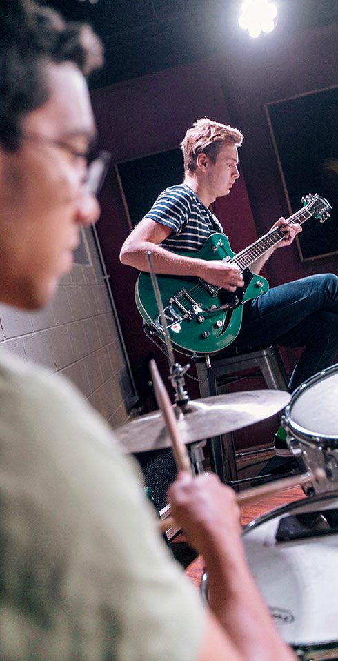 A student in a striped t-shirt plays a green electric guitar sitting on a stool.